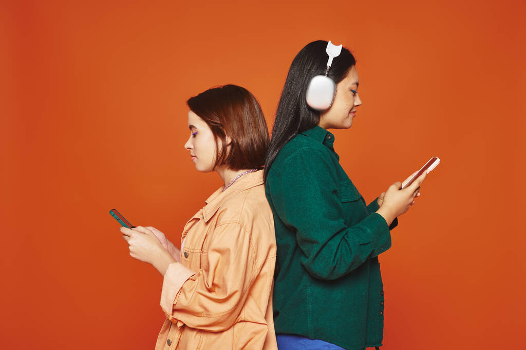onnellinen aasialainen tyttö langattomissa kuulokkeissa pitämällä älypuhelinta lähellä hänen naispuolista ystäväänsä, selkä selkää vasten - Valokuva, kuva