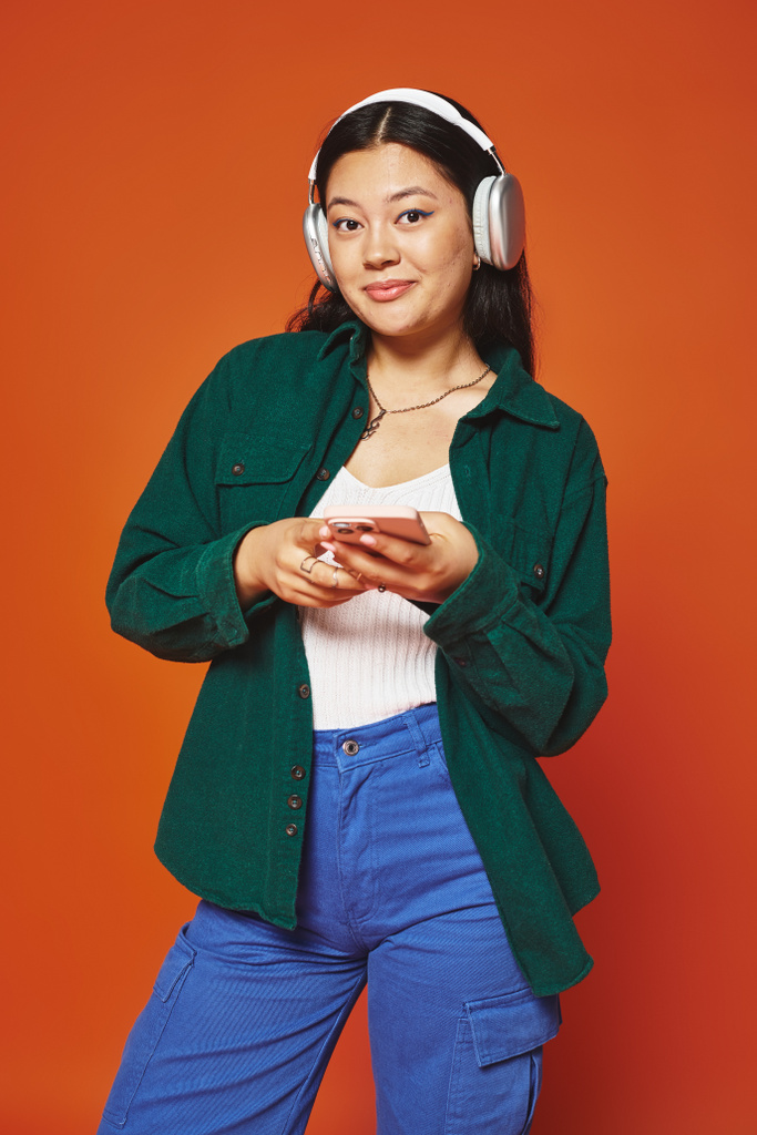 オレンジ色の背景にスマートフォンを保持するワイヤレスヘッドフォンで音楽を聞く陽気なアジアの女性 - 写真・画像