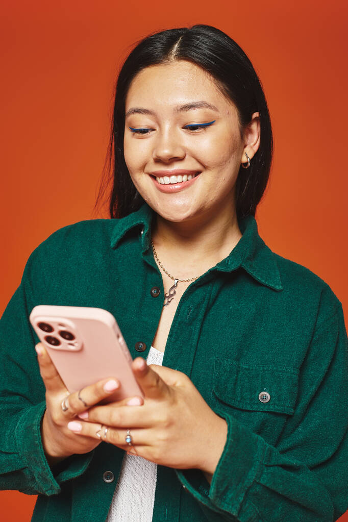 happy and young asian woman with brunette hair using smartphone and smiling on orange background - Photo, Image