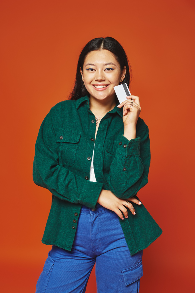 pozytywna młoda azjatycka kobieta w zielonej kurtce trzyma kartę kredytową na pomarańczowym tle, konsumpcjonizm - Zdjęcie, obraz