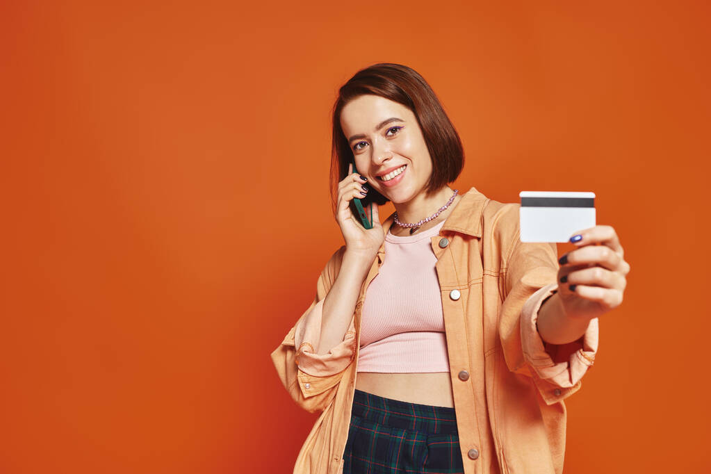 szczęśliwa kobieta rozmawia na smartfonie i trzyma kartę kredytową na pomarańczowym tle, zakupy online - Zdjęcie, obraz