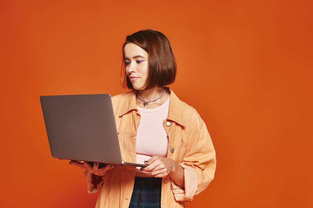 молодой фрилансер в повседневной одежде держит ноутбук и работает удаленно на оранжевом фоне - Фото, изображение