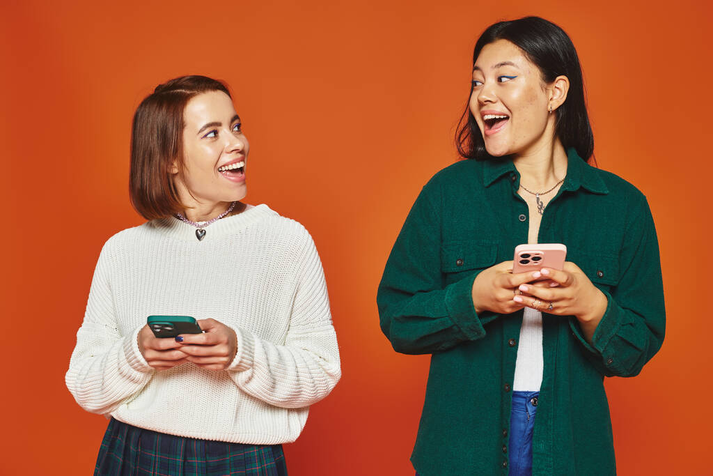 活気に満ちたオレンジ色の背景でスマートフォンを使用する多文化の若い女性,ソーシャルメディア - 写真・画像