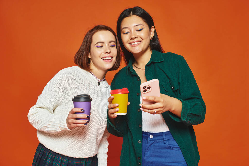 cheerful multicultural female friends holding paper cups with coffee and looking at smartphone - Photo, Image