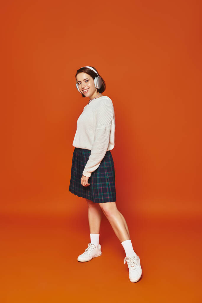 白いセーターとオレンジ色の背景で音楽を聞いているワイヤレスヘッドフォンの肯定的な若い女性 - 写真・画像