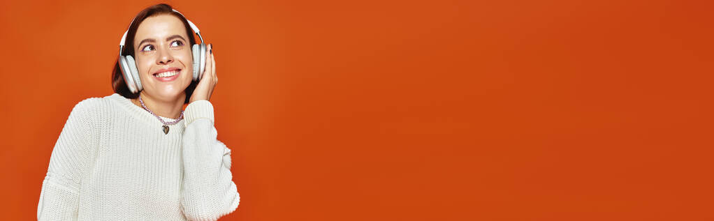 vidám nő fehér pulóverben és vezeték nélküli fejhallgató zenét hallgat narancssárga háttérrel, banner - Fotó, kép