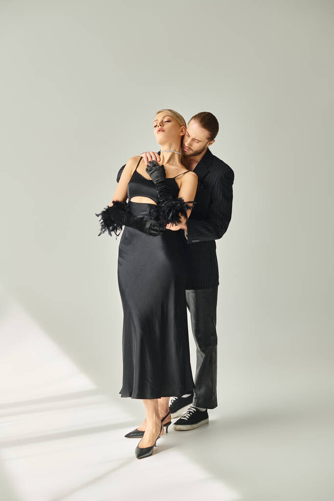 přitažlivý sexy pár v elegantním černém oblečení pózování spolu a objímání láskyplně na šedém pozadí - Fotografie, Obrázek