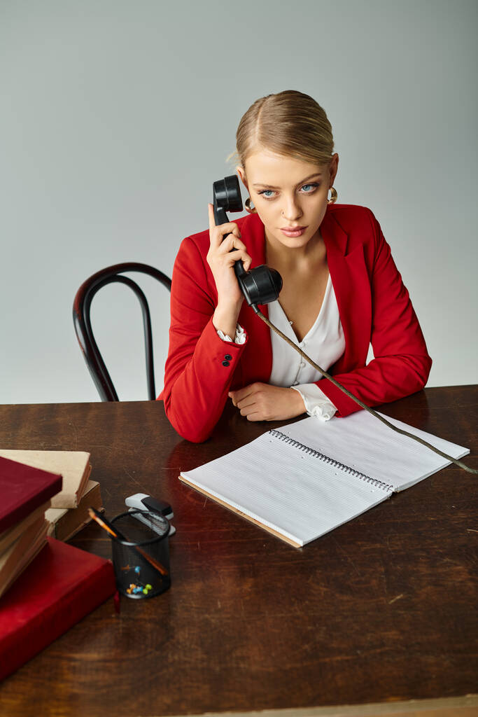 mooie blonde vrouw in rood elegant jasje praten door retro telefoon tijdens het werken op kantoor - Foto, afbeelding