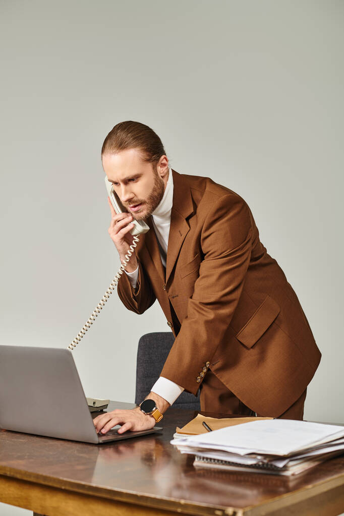 good looking bearded man in stylish suit talking by retro phone and looking at his laptop in office - Photo, Image