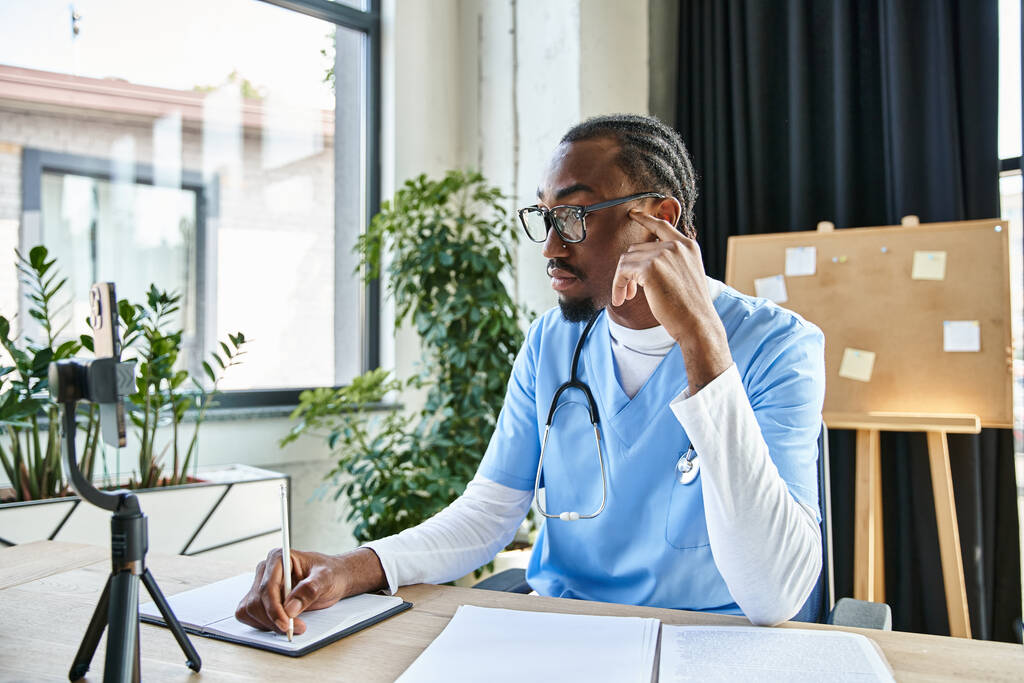 pensive handsome african american doctor taking notes while consulting by phone, telemedicine - Photo, Image