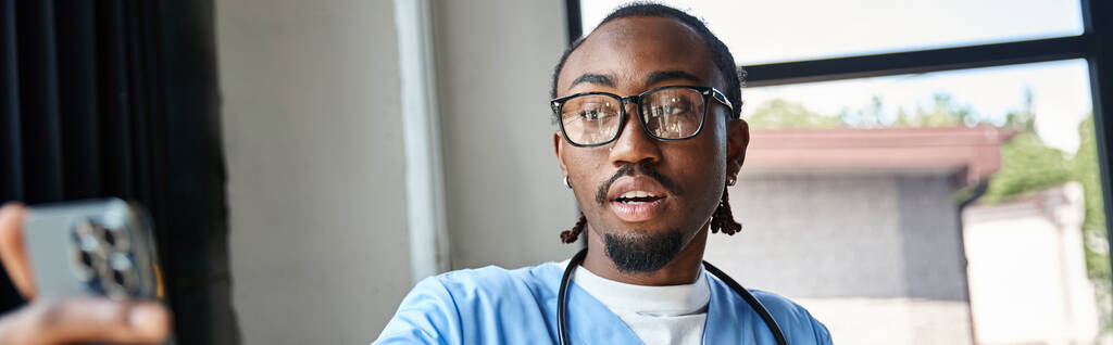 młody, afrykański lekarz w okularach konsultujący się z kimś przez telefon komórkowy, telemedycynę, baner - Zdjęcie, obraz
