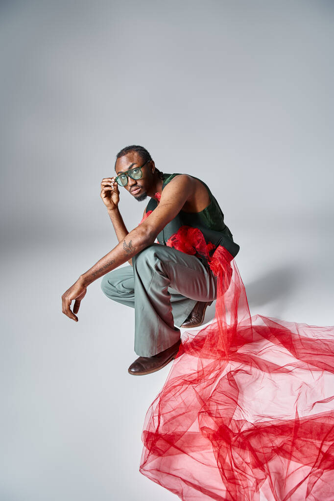 atraente afro-americano homem na moda roupa agachamento no fundo cinza, conceito de moda - Foto, Imagem