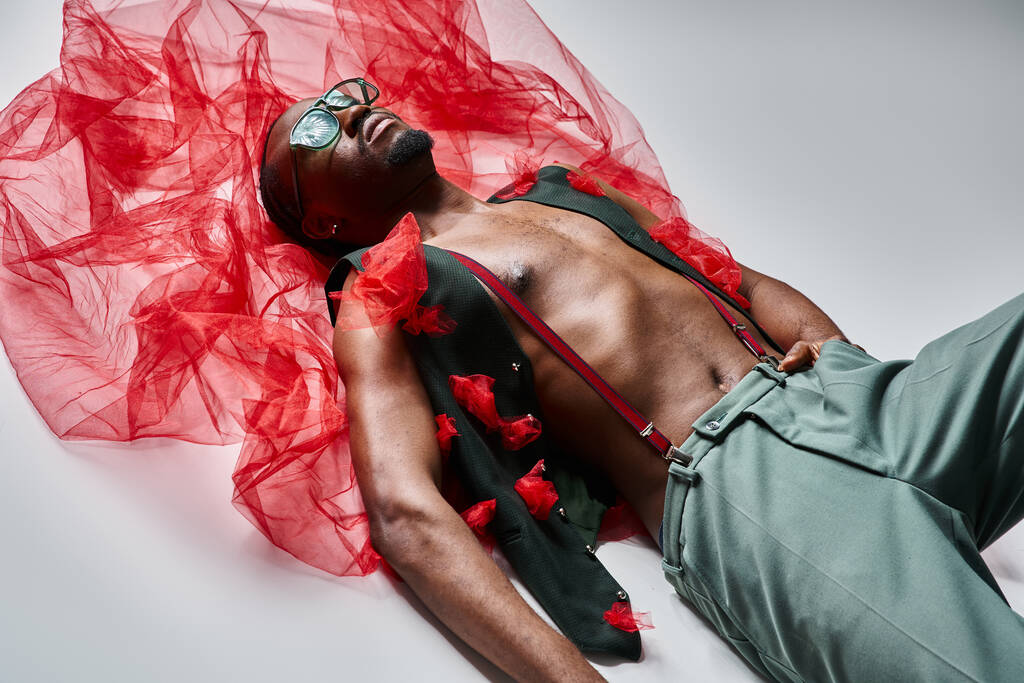 привлекательный молодой африканский мужчина-модель в стильной одежде из красной ткани тюля, лежащей на полу - Фото, изображение