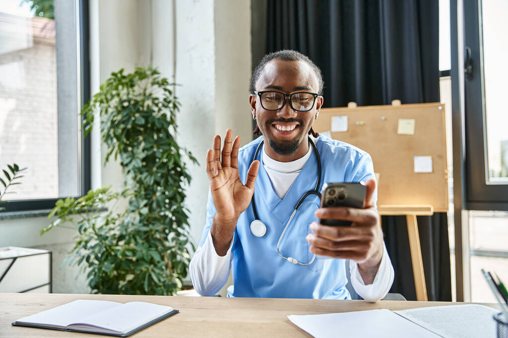 веселий афроамериканський лікар в окулярах махає на камеру мобільного телефону і щасливо посміхається - Фото, зображення