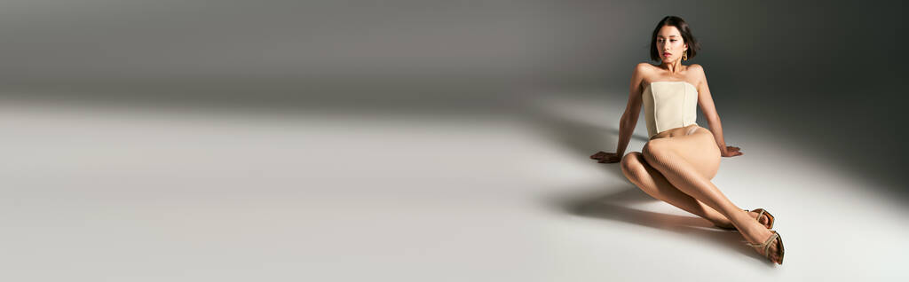 λάβαρο της Ασιάτισσας με κοντά μαλλιά ποζάροντας σε κορσέ και μπεζ καλσόν fishnet σε γκρι φόντο - Φωτογραφία, εικόνα