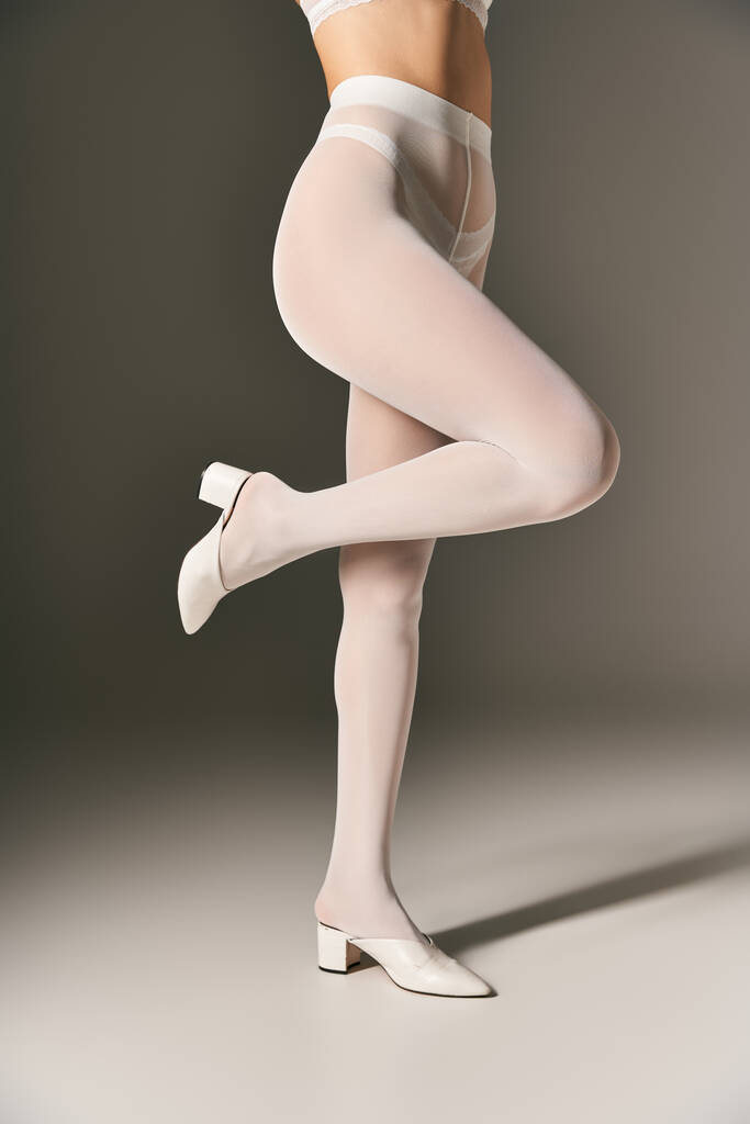 обрезанная фотография стройная женщина в белых колготках и на высоких каблуках позирует на сером фоне - Фото, изображение