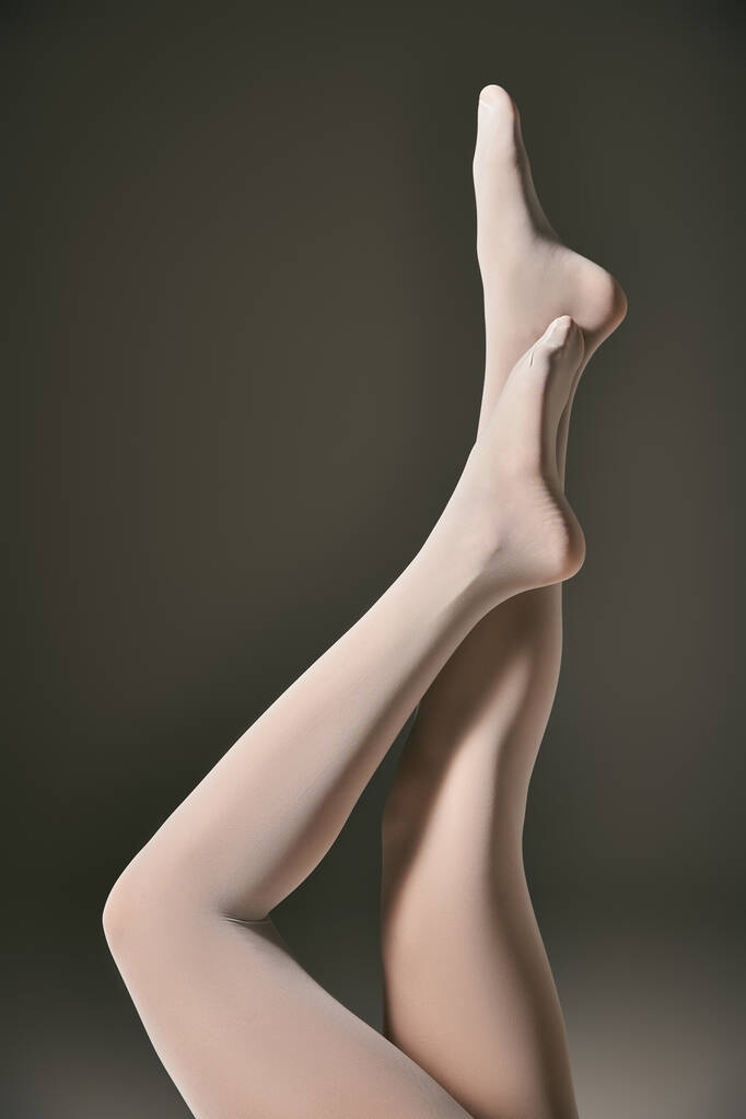 обрізаний вид молодої моделі в білих колготках позує з піднятими ногами на темно-сірому фоні - Фото, зображення