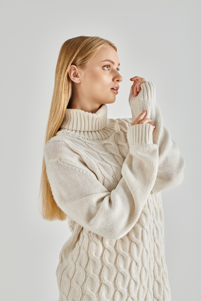 femme blonde gracieuse et réfléchie en pull tricoté blanc regardant loin sur gris, vêtements d'hiver confortables - Photo, image