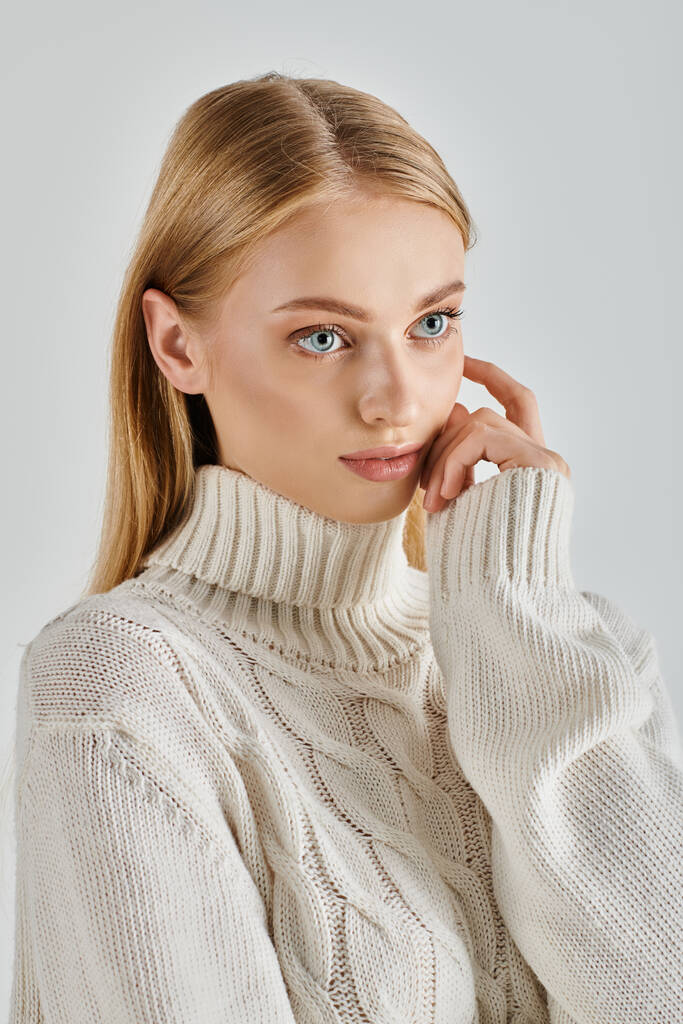 atractiva y soñadora mujer en suéter blanco cálido tocando la cara y mirando hacia otro lado en el fondo gris - Foto, Imagen
