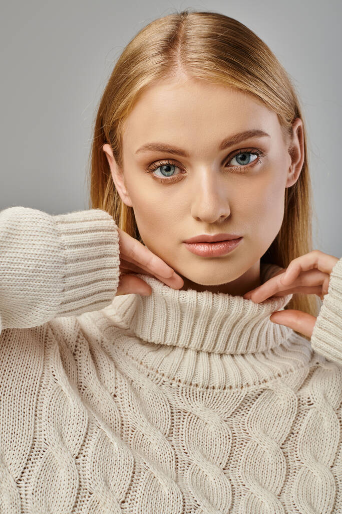 портрет блондинки с натуральным макияжем и выразительным взглядом, позирующим в вязаном свитере на сером - Фото, изображение