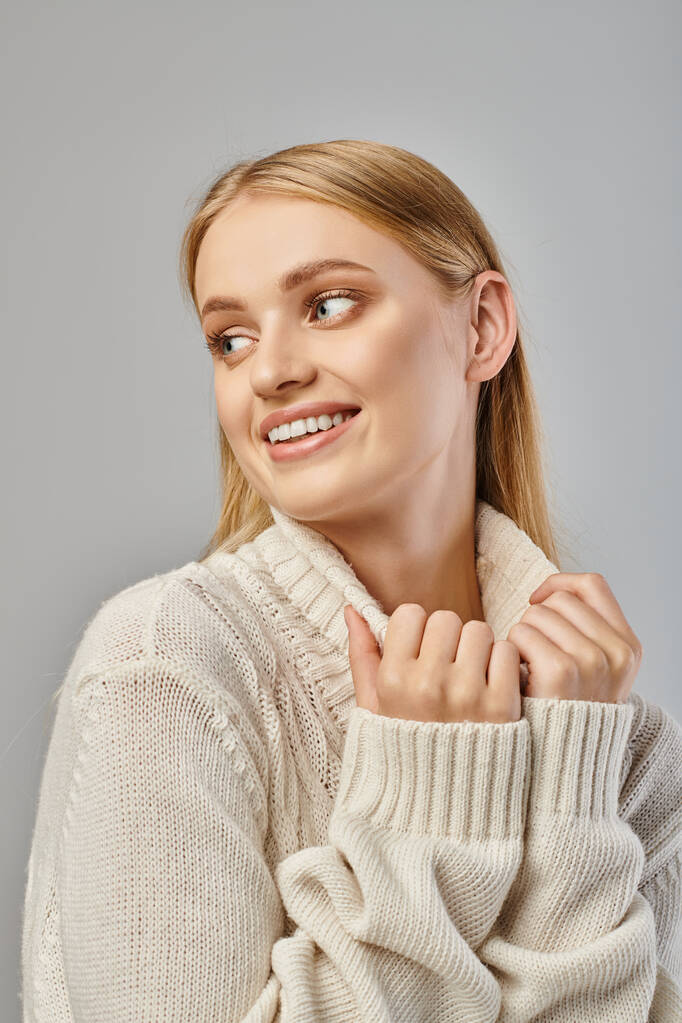 veselá blondýnka v bílém pleteném svetru s úsměvem a pohledem pryč na šedé, zimní štěstí - Fotografie, Obrázek