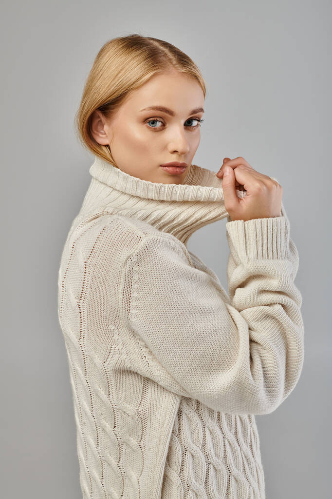 jeune femme blonde en pull tricoté chaud avec col montant regardant la caméra sur gris, tendance hiver - Photo, image