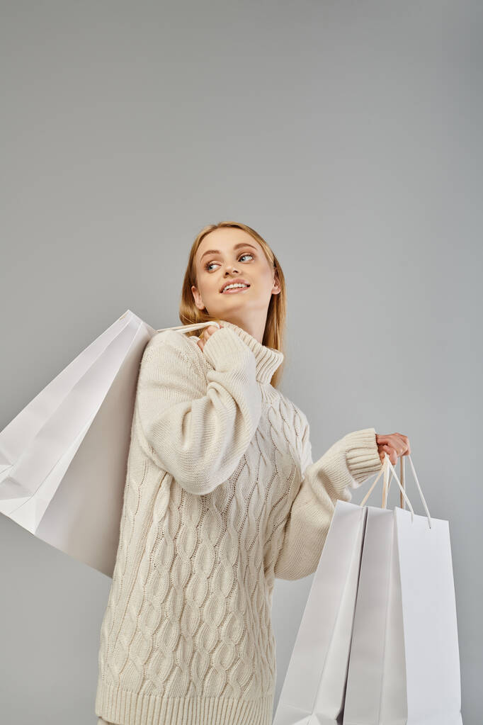 mujer rubia sonriente en suéter de punto suave sosteniendo bolsas de compras blancas y mirando hacia otro lado en gris - Foto, Imagen