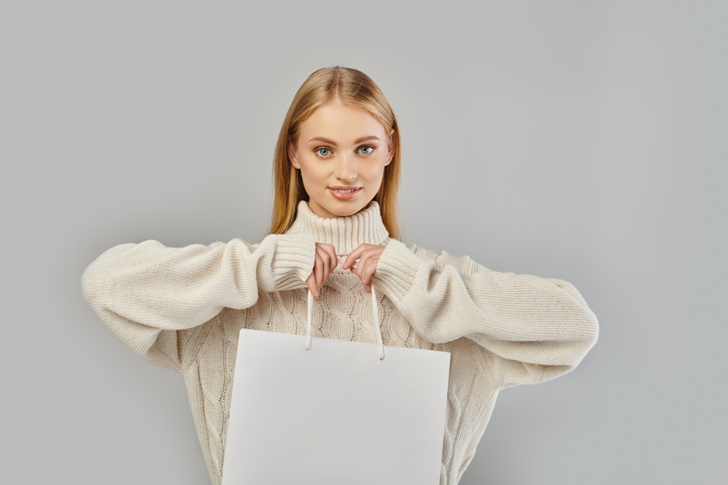 mujer rubia complacida en suéter de punto blanco sosteniendo bolsa de compras y mirando a la cámara en gris - Foto, imagen