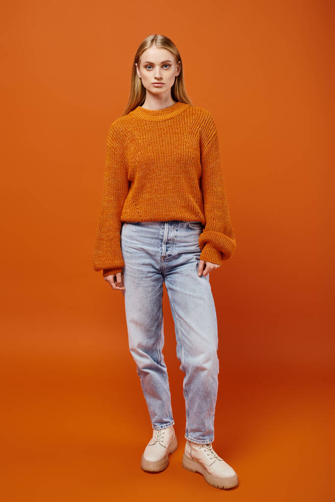 オレンジ色の背景,冬に編まれたセーターとブルージーンズでスタイリッシュなブロンドの女性の全長 - 写真・画像