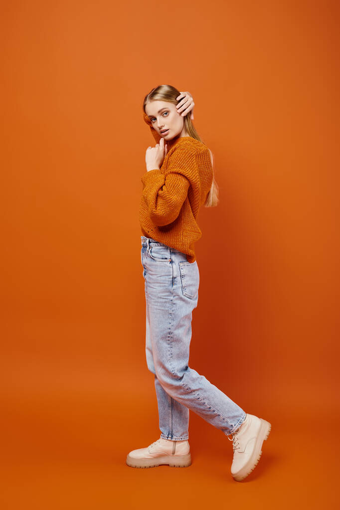 kusząca blondynka w dżinsach i ciepły sweter z dzianiny pozowanie na pomarańczowym tle, trend zimowy - Zdjęcie, obraz