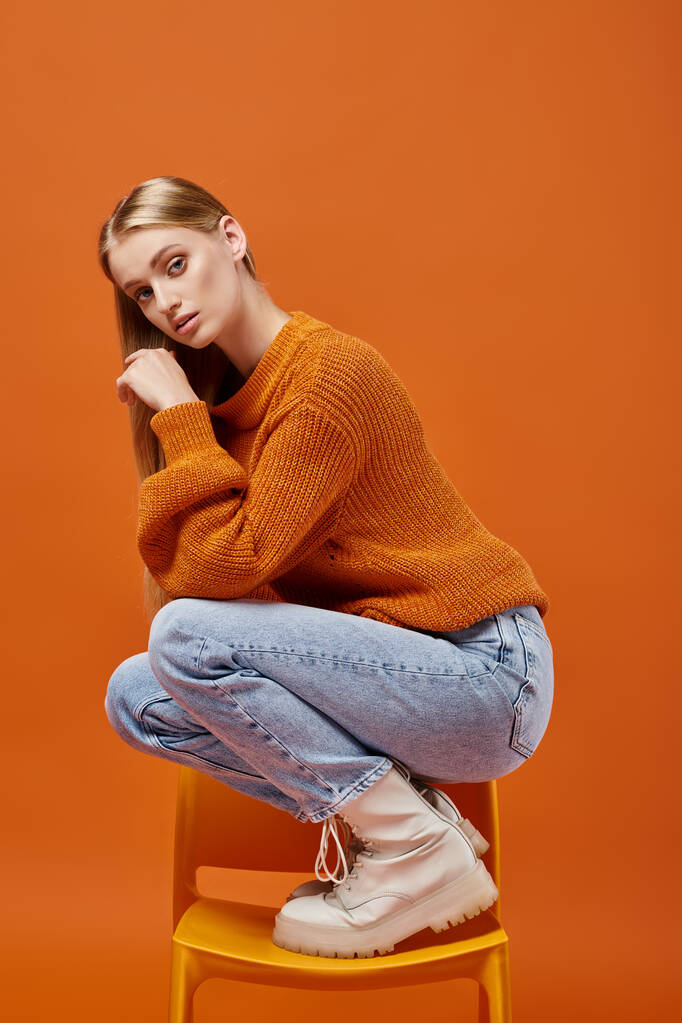 молода блондинка в теплий зимовий светр і джинси позує на стілець і дивиться на камеру на помаранчевому - Фото, зображення