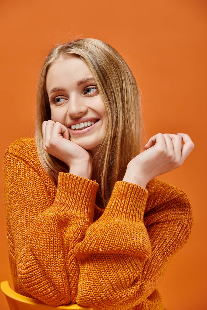 smiley blonde vrouw in helder en gezellig gebreid met natuurlijke make-up kijken naar camera op oranje - Foto, afbeelding