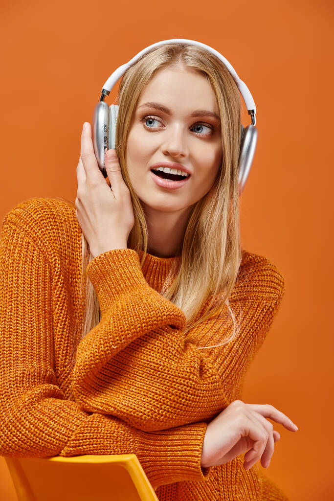 donna stupita in maglione caldo luminoso ascoltare musica in cuffia su arancione, emozioni invernali - Foto, immagini