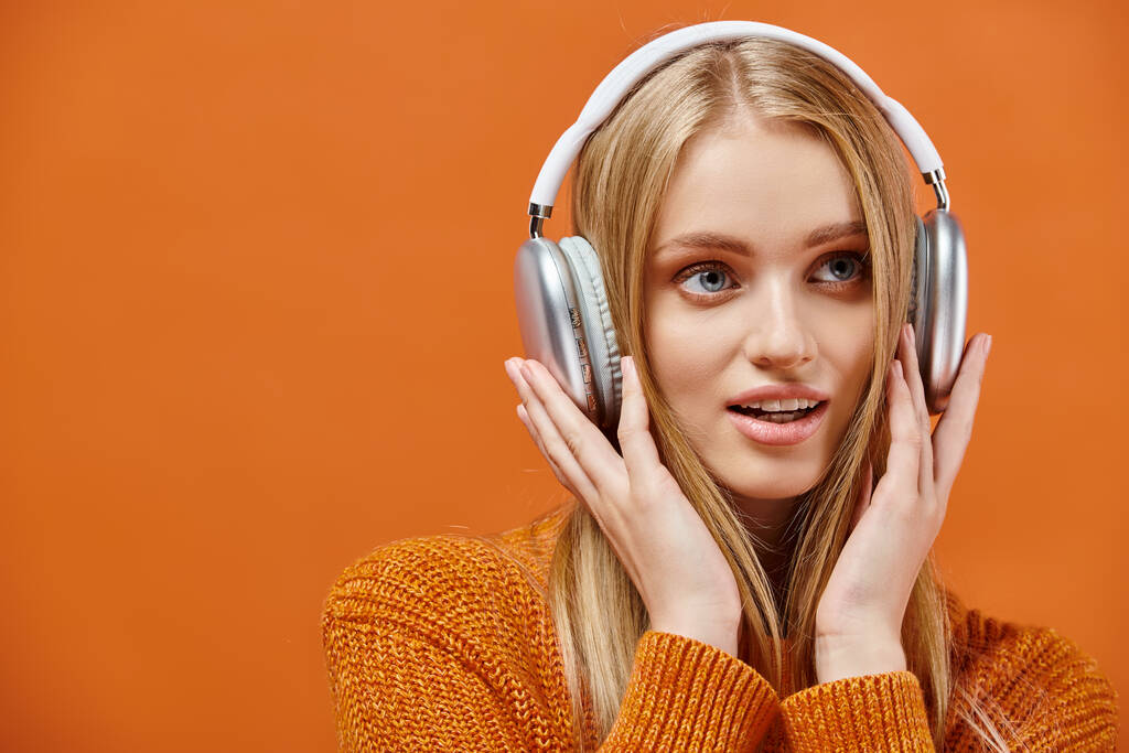 impressed woman in warm knitted sweater listening music in headphones on orange, winter emotions - Photo, Image