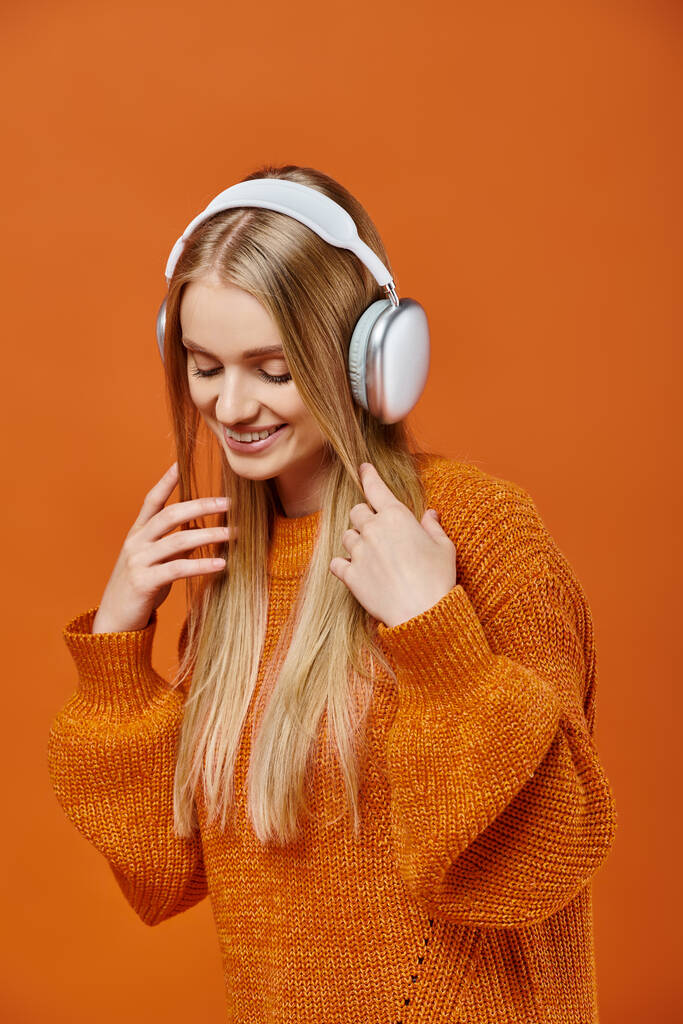 smiling blonde woman in warm orange sweater and headphones listening music on bright backdrop - Photo, Image