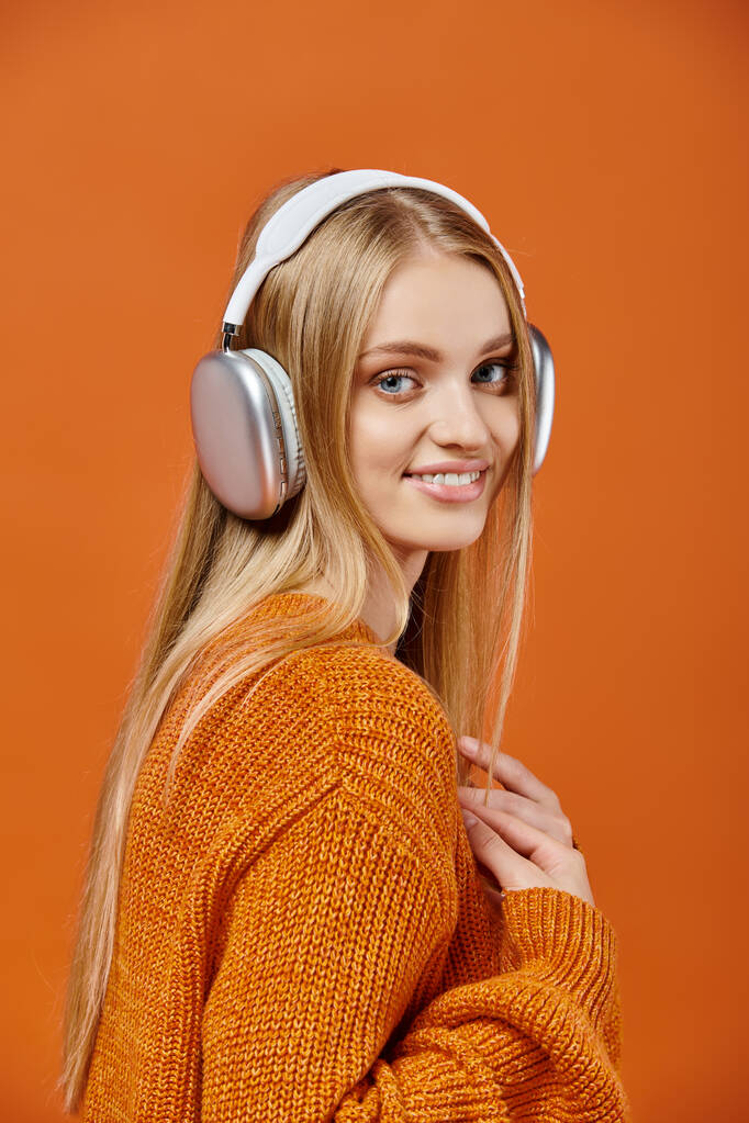 jovem mulher alegre em camisola de inverno laranja e fones de ouvido ouvindo música e sorrindo para a câmera - Foto, Imagem
