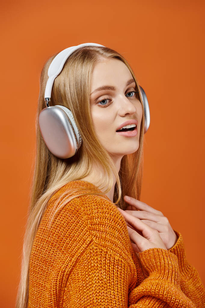 Parlak süveter ve kulaklıklar takan, müzik dinleyen ve portakala bakan sarışın bir kadın. - Fotoğraf, Görsel