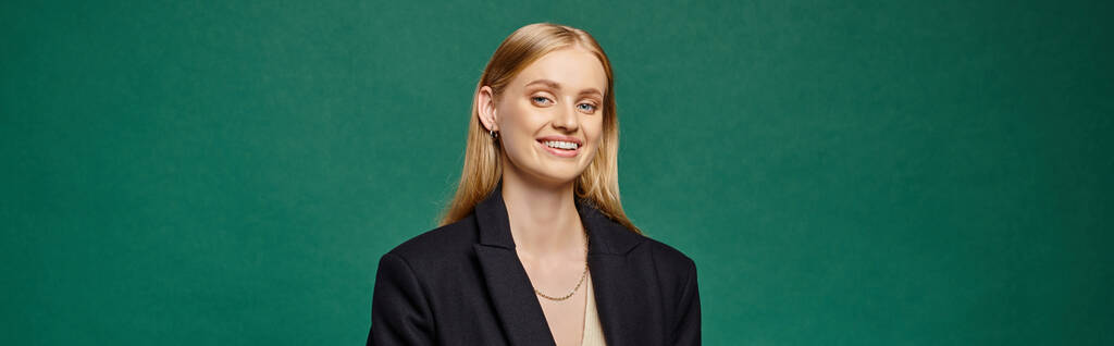 joyful and stylish blonde woman in black warm coat looking at camera on green, horizontal banner - Photo, Image