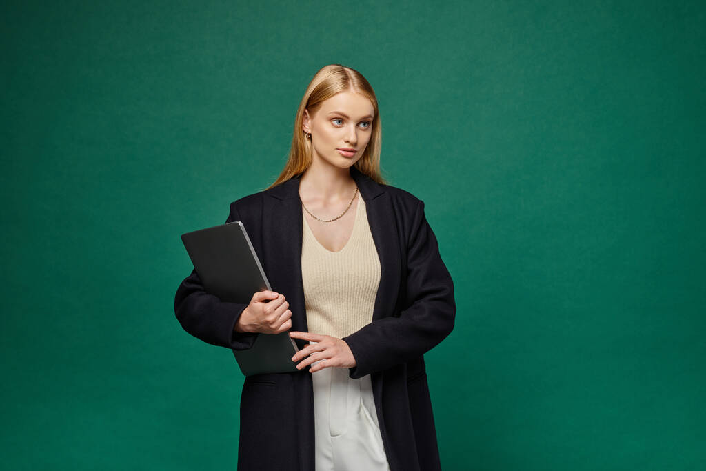 модная блондинка в черном зимнем пальто держит ноутбук и смотрит в сторону на зеленом фоне - Фото, изображение