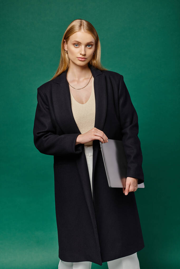young blonde woman in black winter coat holding laptop and looking at camera on green backdrop - Photo, Image