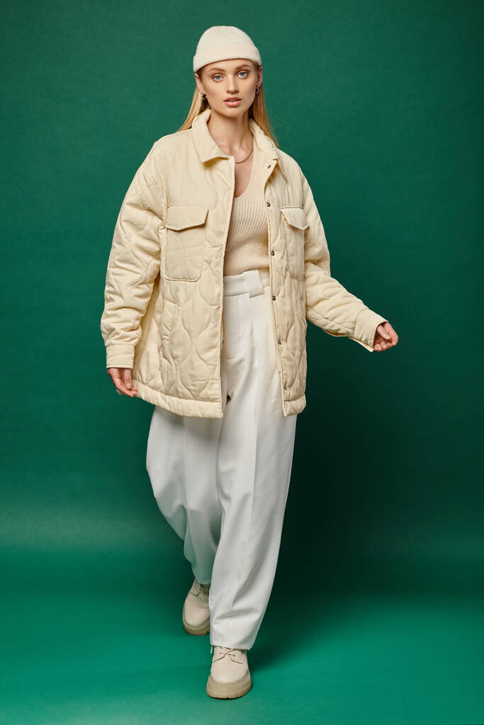 okouzlující a trendy ženský model v slonovinové bundě a čepici kráčející po zelené, zimní módě - Fotografie, Obrázek