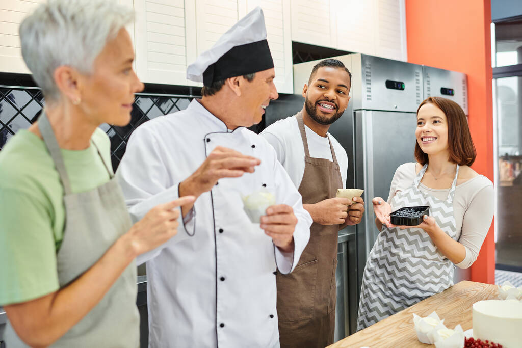 gente multicultural alegre en delantales hablando con cocinero maduro en sombrero blanco, cursos de cocina - Foto, imagen