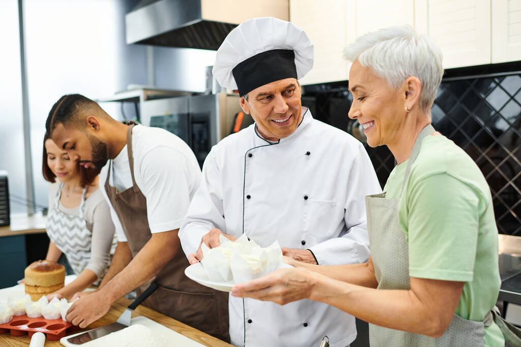 ώριμος χαρούμενος σεφ σε λευκό καπέλο διδασκαλία πώς να ψήνουν διαφυλετικούς μαθητές του κατά τη διάρκεια του μαθήματος μαγειρικής - Φωτογραφία, εικόνα