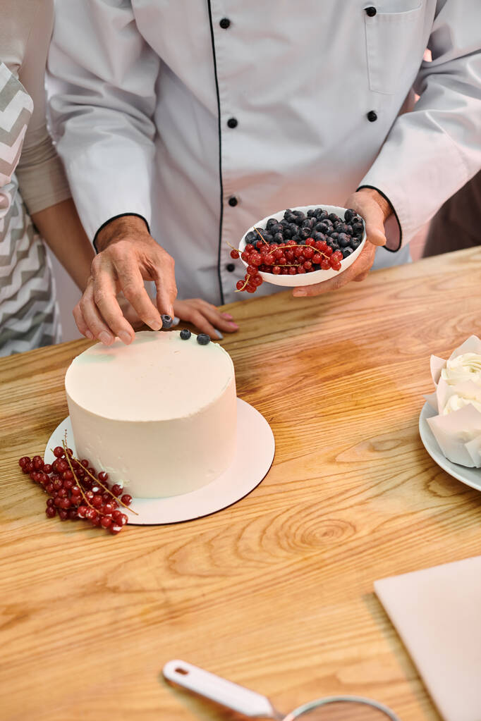 rajattu näkymä kypsä kokki opettaa nuorelle opiskelijalle, miten koristella kakku marjoilla oppitunnilla - Valokuva, kuva