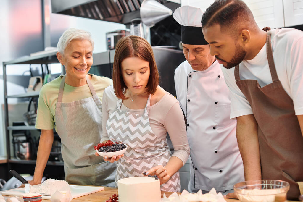 jonge vrouw met schort versieren taart naast haar multiraciale vrolijke vrienden en chef-kok in witte hoed - Foto, afbeelding