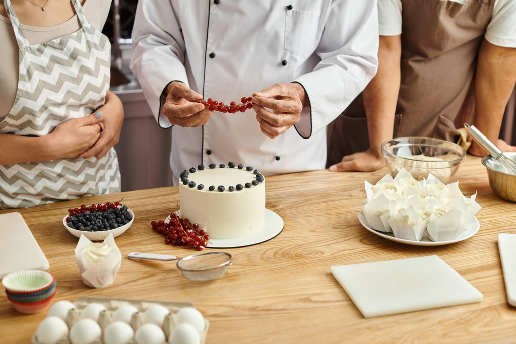 vista recortada de chef maduro decorando delicioso pastel con grosella roja junto a sus diversos estudiantes - Foto, imagen