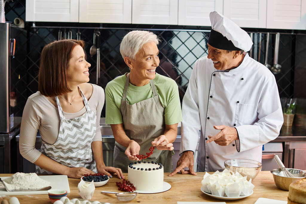 freudige junge Frau in Schürze lächelt Koch und ihren reifen Freund an, der Kuchen mit Beeren dekoriert - Foto, Bild