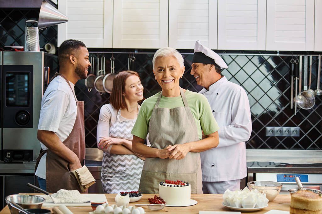zaměření na zralou ženu s dort úsměv na kameru s rozmazané rozmanité přátele a kuchař na pozadí - Fotografie, Obrázek