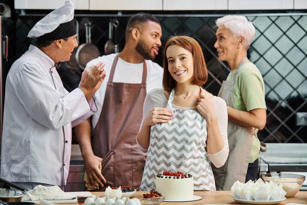 νεαρή χαρούμενη γυναίκα με ποδιά κοιτάζοντας την κάμερα δίπλα στην τούρτα με τους φίλους της στο σκηνικό - Φωτογραφία, εικόνα