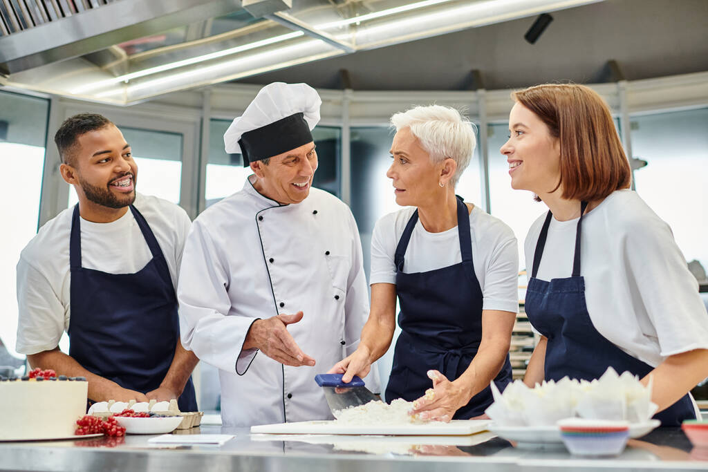 mulher bonita madura no avental trabalhando com massa e olhando para chefs interracial alegres - Foto, Imagem